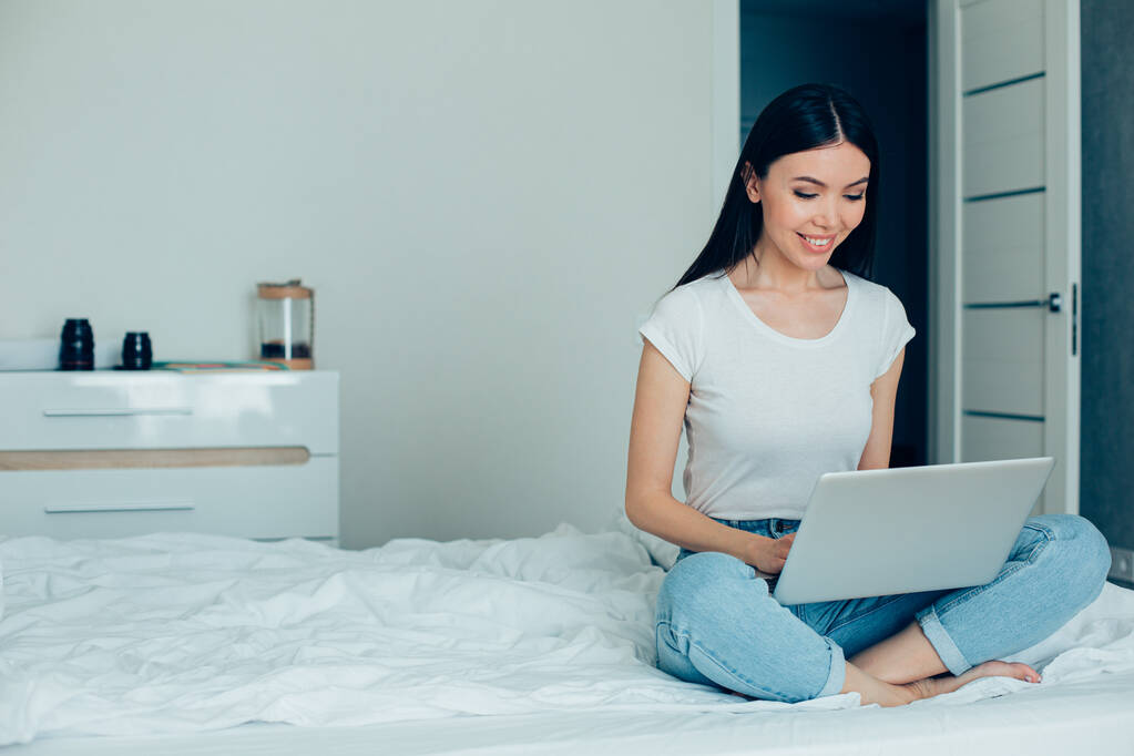Χαρούμενη χαμογελαστή κυρία που κάθεται στο κρεβάτι με ένα μοντέρνο φορητό υπολογιστή στην αγκαλιά της και το χρησιμοποιεί. Πανό ιστοχώρου - Φωτογραφία, εικόνα