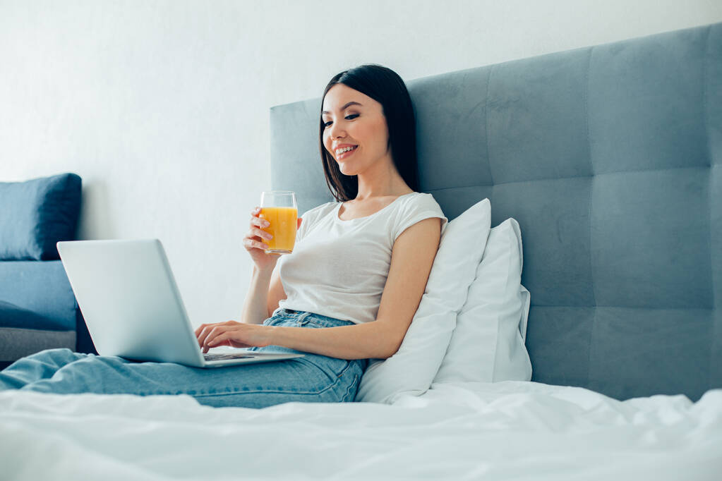 Mooie jonge blanke vrouw zittend in de slaapkamer met een glas sinaasappelsap en kijkend naar het laptopscherm - Foto, afbeelding