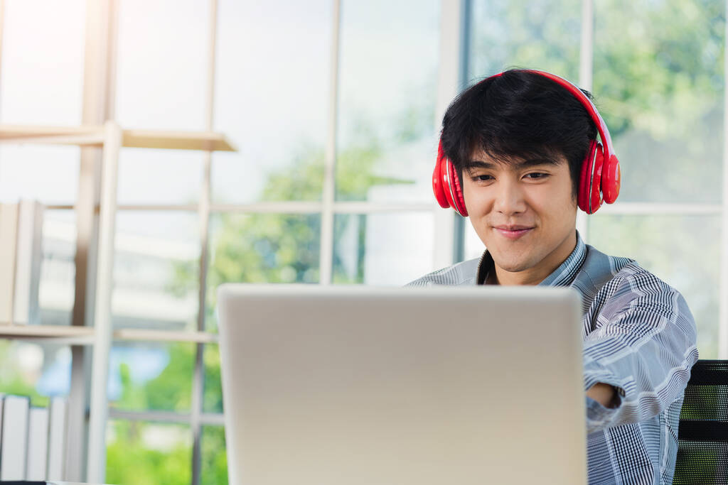 Азиатский молодой бизнесмен счастье с красными наушниками он сидит на рабочем столе домашний офис ищет ноутбук монитор компьютера, уверенный красивый мужчина образ жизни улыбка расслабляет он слушает музыку - Фото, изображение