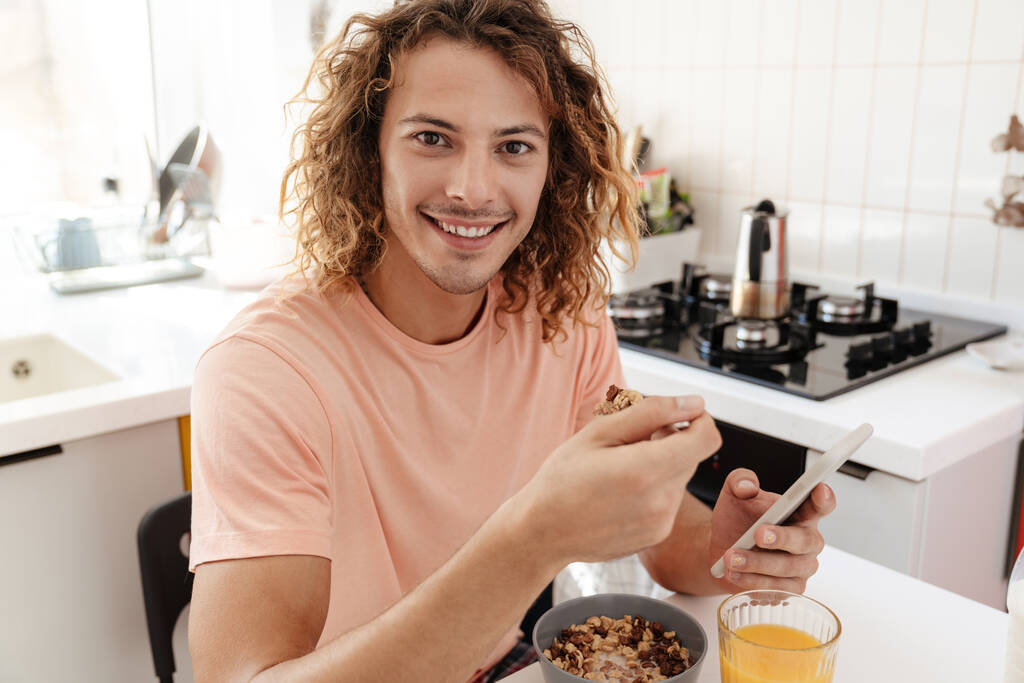 Caucasiano bonito sorrindo cara usando celular enquanto toma café da manhã na cozinha - Foto, Imagem