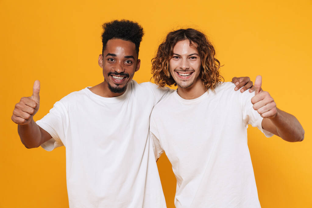 Happy multicultural two guys showing thumbs up and hugging isolated over yellow background - Photo, Image