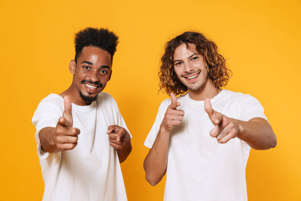 Happy multicultural two guys smiling and pointing fingers at camera isolated over yellow background - 写真・画像