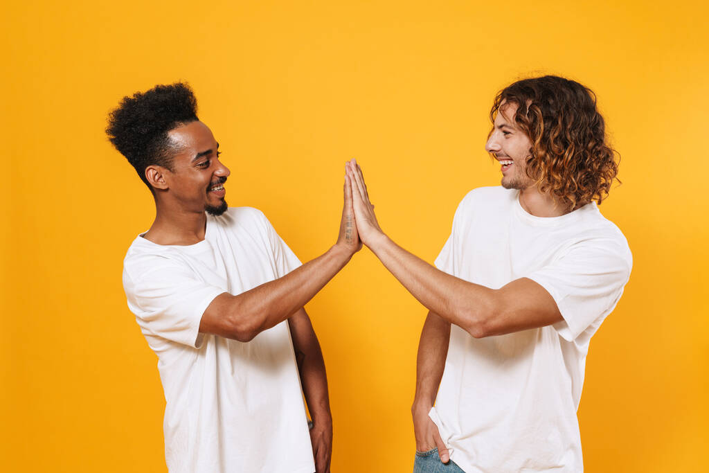 Happy multicultural two guys smiling and giving high five isolated over yellow background - Photo, Image