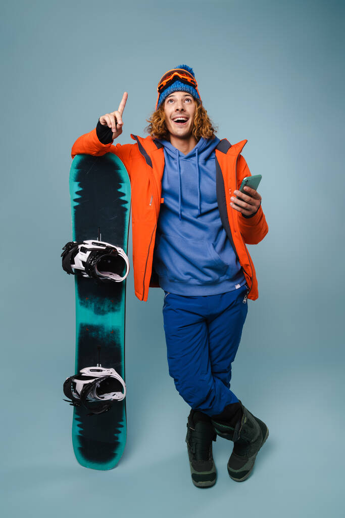 Fešák šťastný sportovec pózující s mobilním telefonem a snowboardem izolované přes modré pozadí - Fotografie, Obrázek