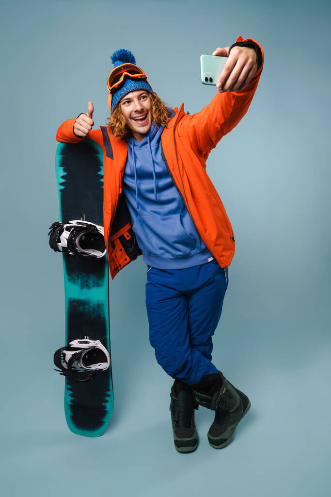 Красивый счастливый спортсмен делает селфи со сноубордом на мобильном телефоне изолированы на синем фоне - Фото, изображение