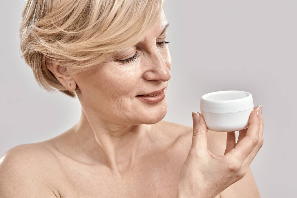 Close-up portret van mooie vrouw van middelbare leeftijd houden, kijken naar hydraterende crème pot terwijl poseren geïsoleerd over grijze achtergrond - Foto, afbeelding