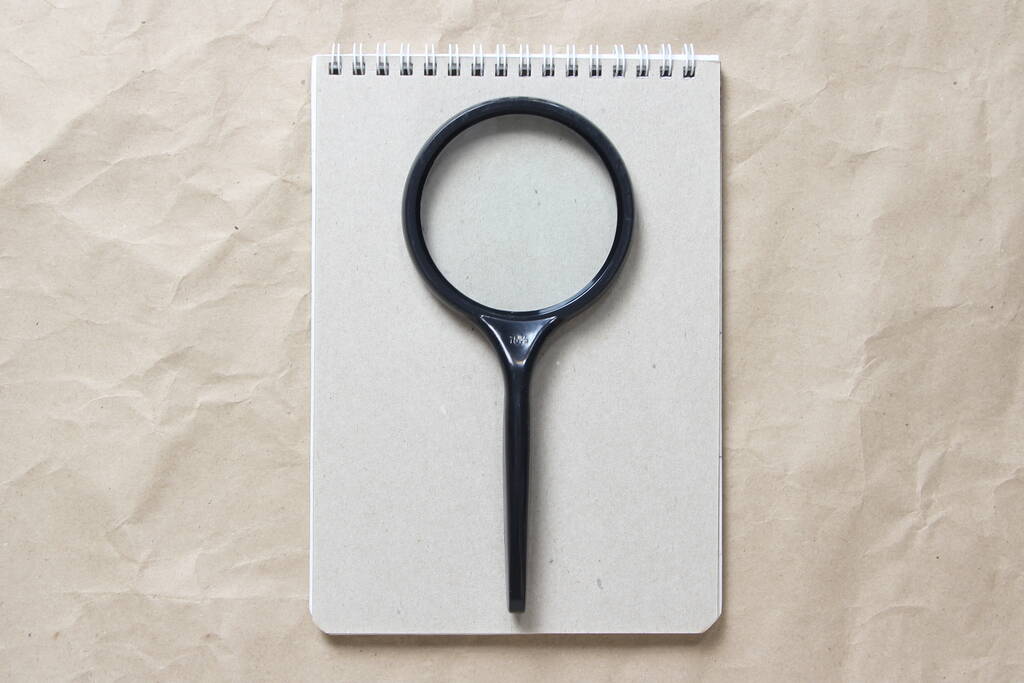 Szary notatnik z białą zwiniętą sprężyną i lupą na tle beżowego pogniecionego papieru. Z pustym miejscem na tekst i projekt - Zdjęcie, obraz