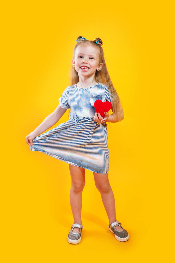 Adorable modelo pequeño en vestido de moda posando con corazón rojo sobre fondo amarillo - Foto, imagen
