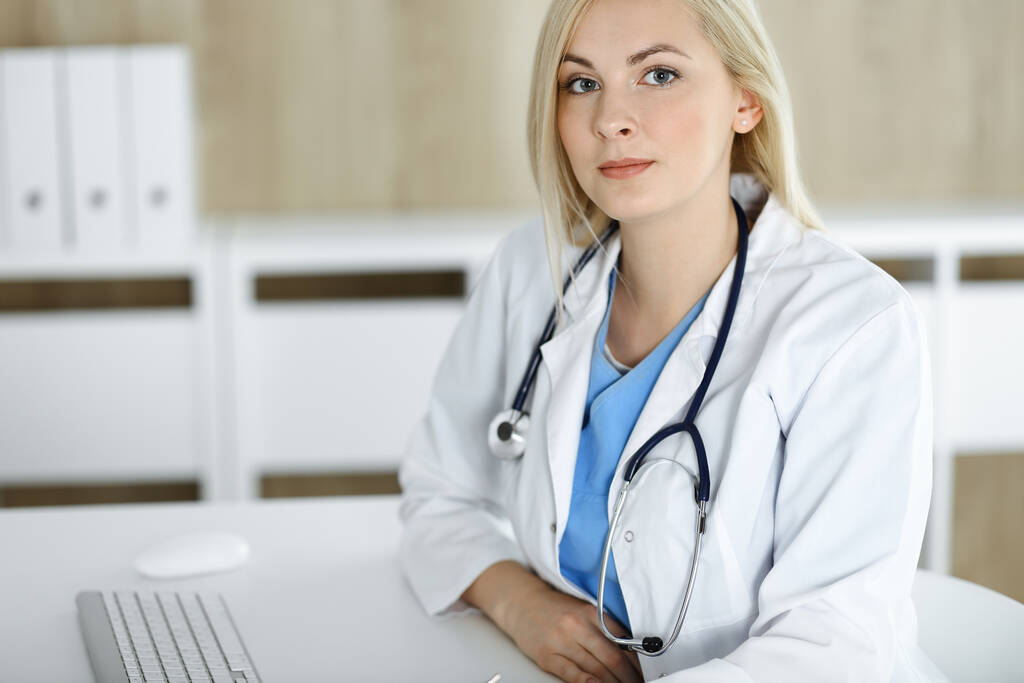 Portret kobiety-lekarza w pracy, siedzącej przy biurku w klinice. Blondynka wesoły lekarz gotowy pomóc pacjentom - Zdjęcie, obraz