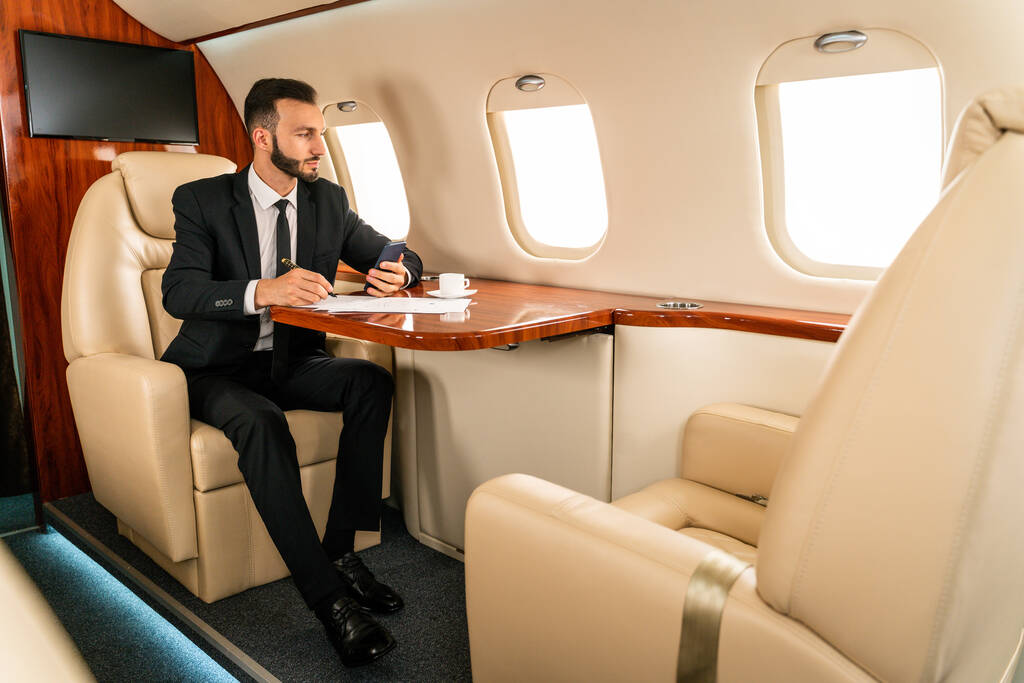 Przystojny biznesmen w eleganckim garniturze latający ekskluzywnym prywatnym odrzutowcem - odnoszący sukcesy przedsiębiorca siedzący w ekskluzywnej klasie biznesowej w samolocie, koncepcje biznesu i trasportacji - Zdjęcie, obraz