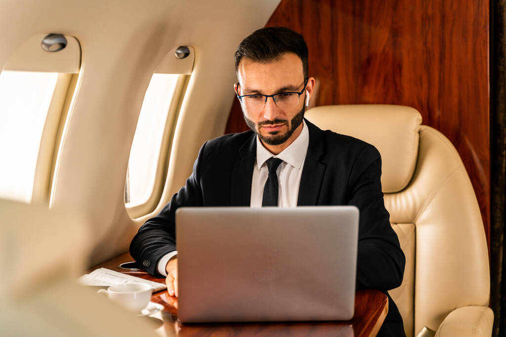 プライベートジェットでエレガントなスーツを着たハンサムなビジネスマン-飛行機の排他的なビジネスクラスに座って成功した起業家、ビジネスと交通に関する概念 - 写真・画像