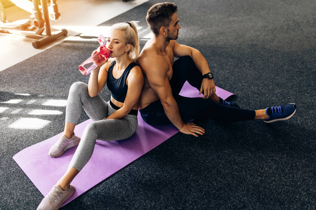 jeune couple sportif en vêtements de sport assis sur des tapis de yoga et tenant des bouteilles d'eau après l'entraînement à la salle de gym - Photo, image