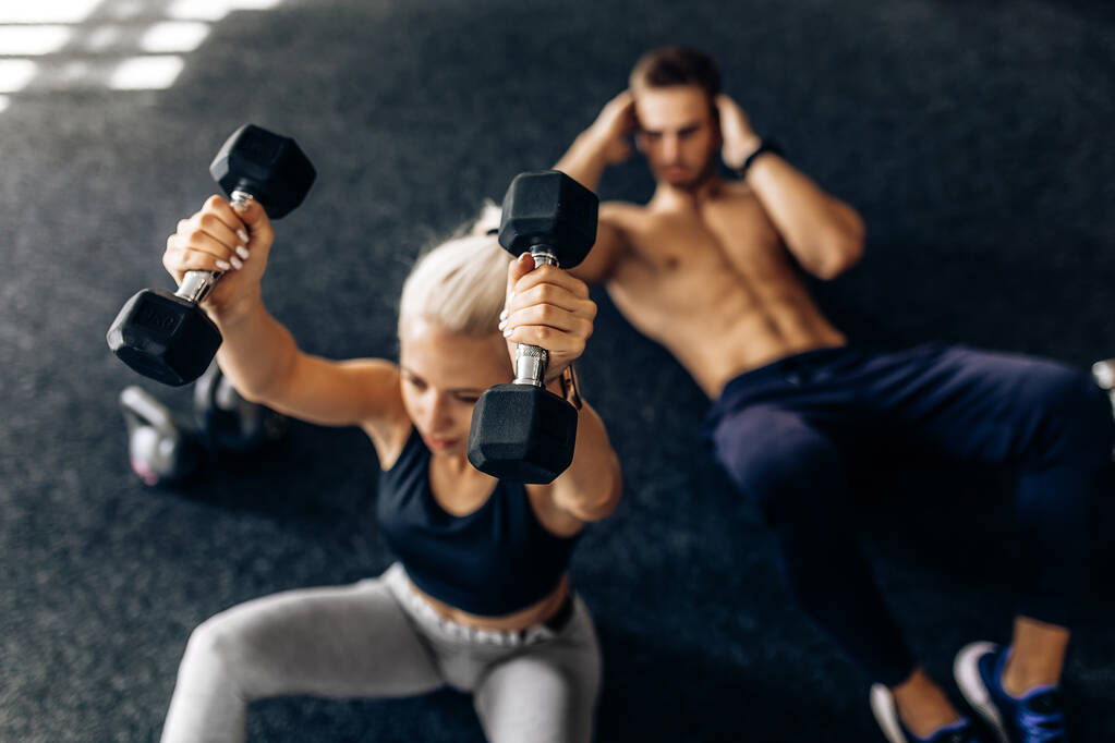 Спортивная молодая пара, мужчина и женщина делают упражнения на животе с гантелями, на полу в тренажерном зале - Фото, изображение