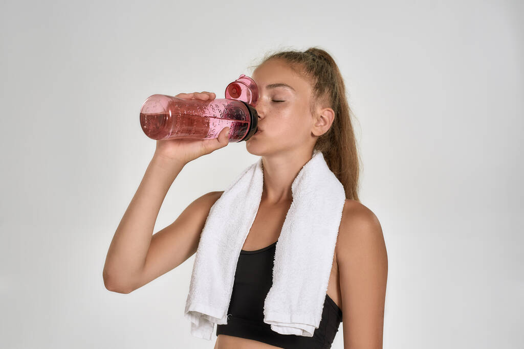 Portret van een schattig sportief meisje kind in sportkleding drinkwater uit de fles terwijl staande met handdoek rond haar nek geïsoleerd over witte achtergrond - Foto, afbeelding