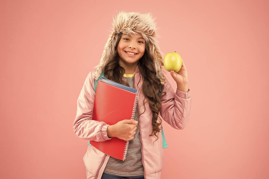 schoolmeisje met boeken na de les. schoolvakantie. klein gelukkig meisje oorflap hoed eet appel. wintervakantie. tiener kind roze achtergrond. pupil dagelijks leven. Modern onderwijs. Student van de maand - Foto, afbeelding