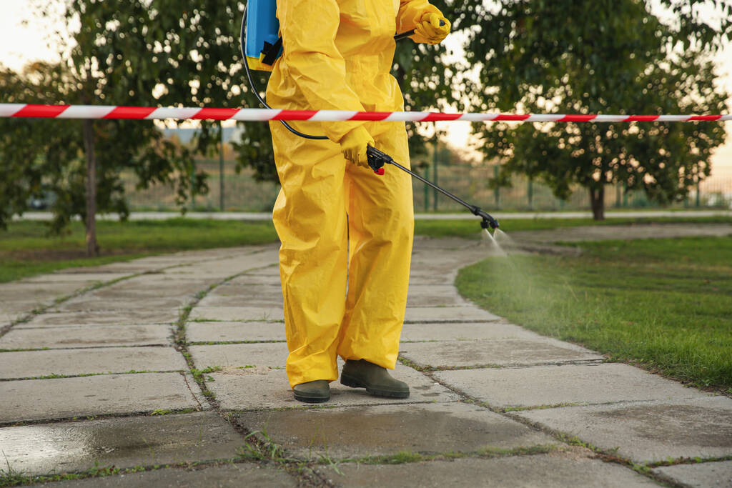 Άτομο με στολή Hazmat που απολυμαίνει οδόστρωμα με ψεκαστήρα, κοντινό πλάνο. Επεξεργασία επιφάνειας κατά τη διάρκεια πανδημίας του κορωναϊού - Φωτογραφία, εικόνα