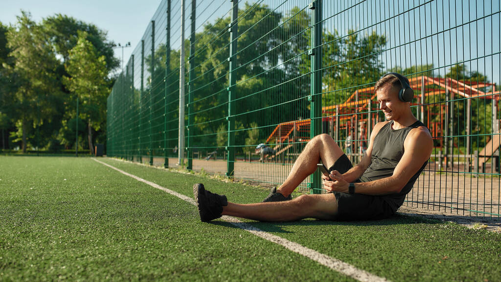 Młody sportowiec w słuchawkach siedzący na boisku, słuchający muzyki, przeglądający playlistę na smartfonie i uśmiechnięty - Zdjęcie, obraz
