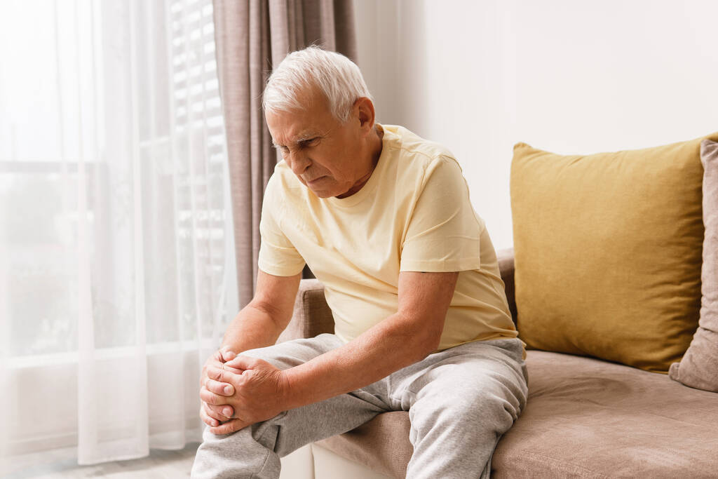 Oudere man die thuis pijn in zijn knie heeft. Concept van problemen met gewrichten zoals artritis. - Foto, afbeelding