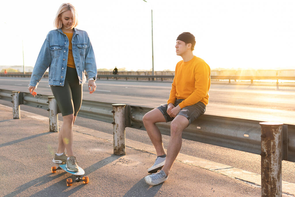 Günbatımında köprüde uzun bir sörf tahtası olan şık genç bir çift. - Fotoğraf, Görsel