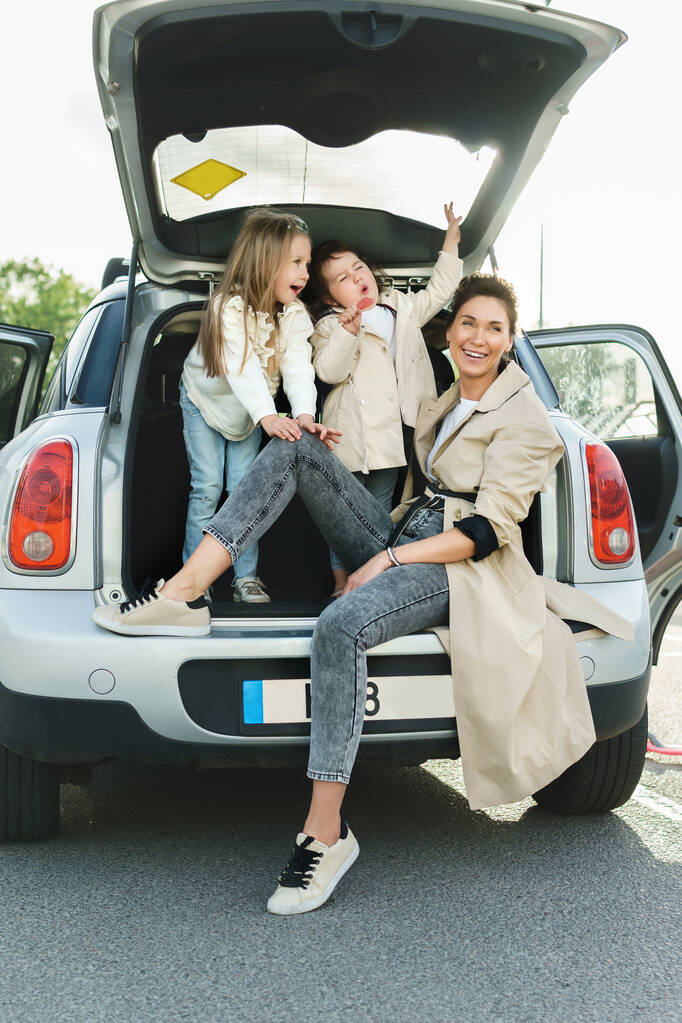 Ευτυχισμένη οικογένεια έτοιμη για ένα οδικό ταξίδι. Η νεαρή μητέρα και οι αξιολάτρευτες κόρες της να κάθονται σε ένα πορτ-μπαγκάζ.. - Φωτογραφία, εικόνα