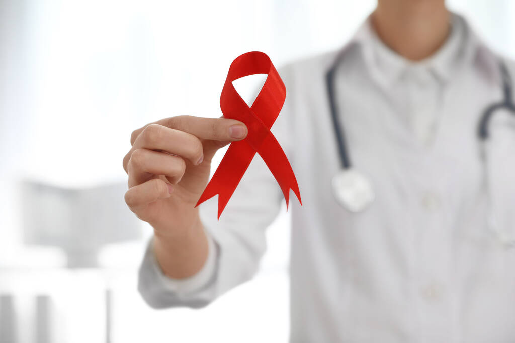Dokter met rood lint op wazige achtergrond, close-up. Aids bewustmakingsmaand - Foto, afbeelding