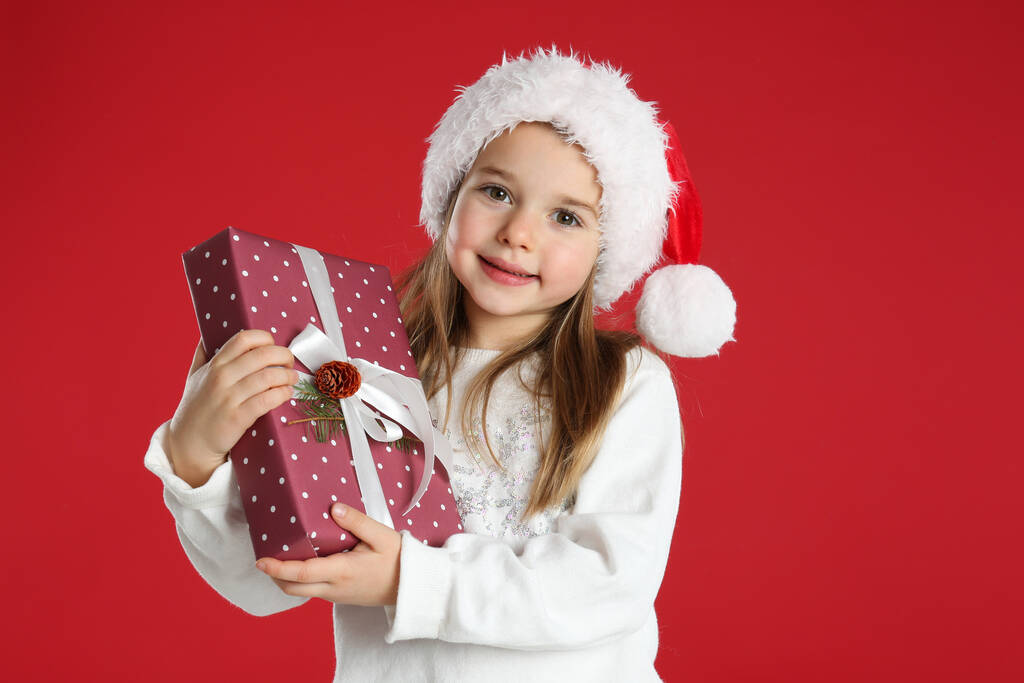 Niedliches Kind in Weihnachtsmütze mit Weihnachtsgeschenk auf rotem Hintergrund - Foto, Bild