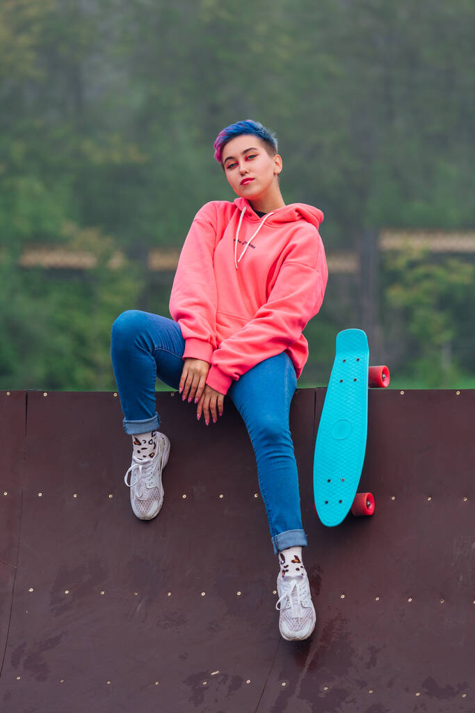 Ritratto di una graziosa ragazza alla moda con capelli corti colorati e piercing al naso vestita con cappuccio rosa e jeans seduta accanto al campo da skateboard con il suo skateboard di plastica blu. - Foto, immagini