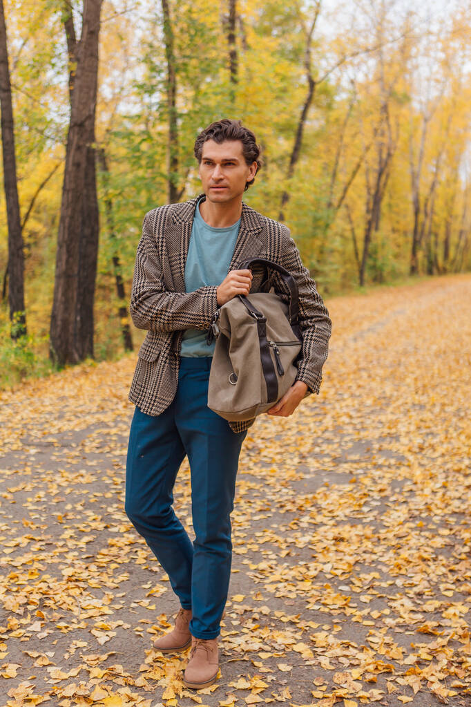 Ψηλός όμορφος άντρας ντυμένος με καφέ μπουφάν περπατώντας με μια τσάντα στο φθινοπωρινό σοκάκι - Φωτογραφία, εικόνα
