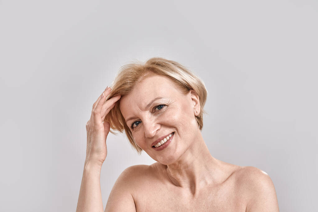 Kaunis keski-ikäinen nainen hymyilee kameralle, säätää hiuksiaan, poseeraa harmaata taustaa vasten. Kauneus, ihonhoito käsite - Valokuva, kuva