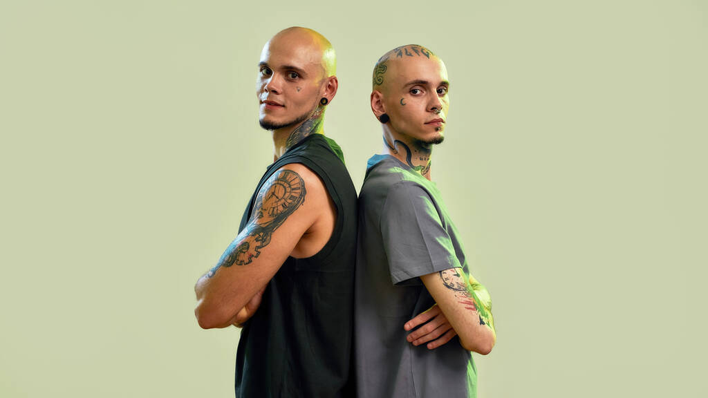 Portrait of two young men, twin brothers with tattoos and piercings looking at camera, posing together, standing back to back isolated over light green background - Photo, Image