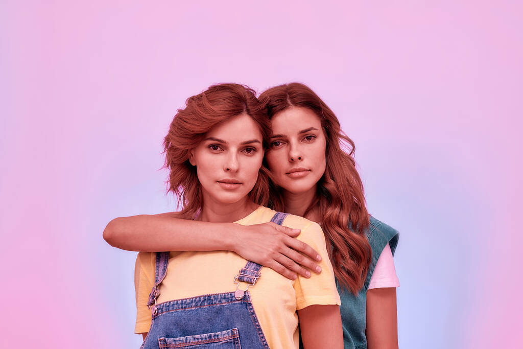 Portrét dvou atraktivních mladých dívek, dvojčata se dívají do kamery, pózují spolu izolovaně přes růžové pozadí - Fotografie, Obrázek