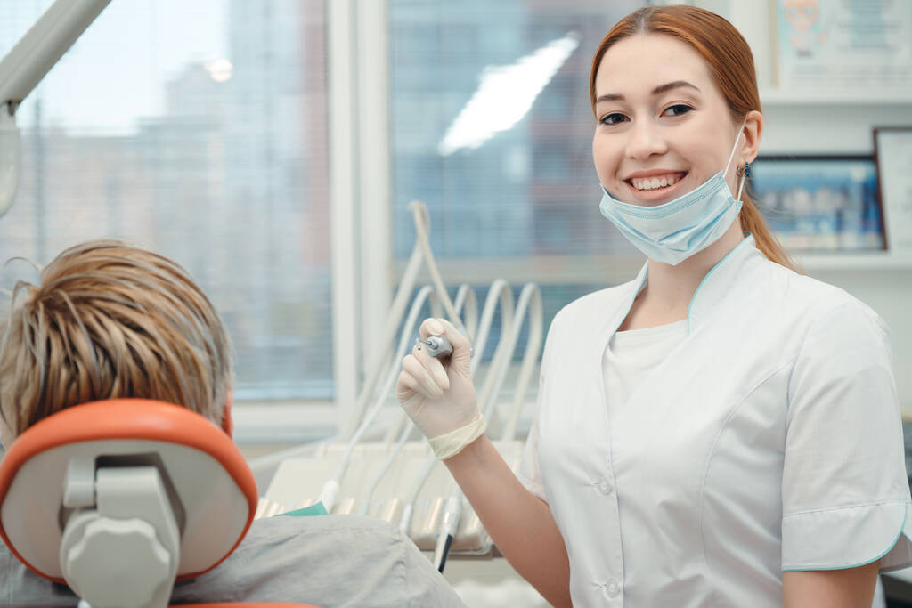 Aantrekkelijke vrouwelijke tandarts in een witte medische jurk met een tandheelkundig instrument in de hand poserend voor de camera naast de patiënt in een stoel - Foto, afbeelding