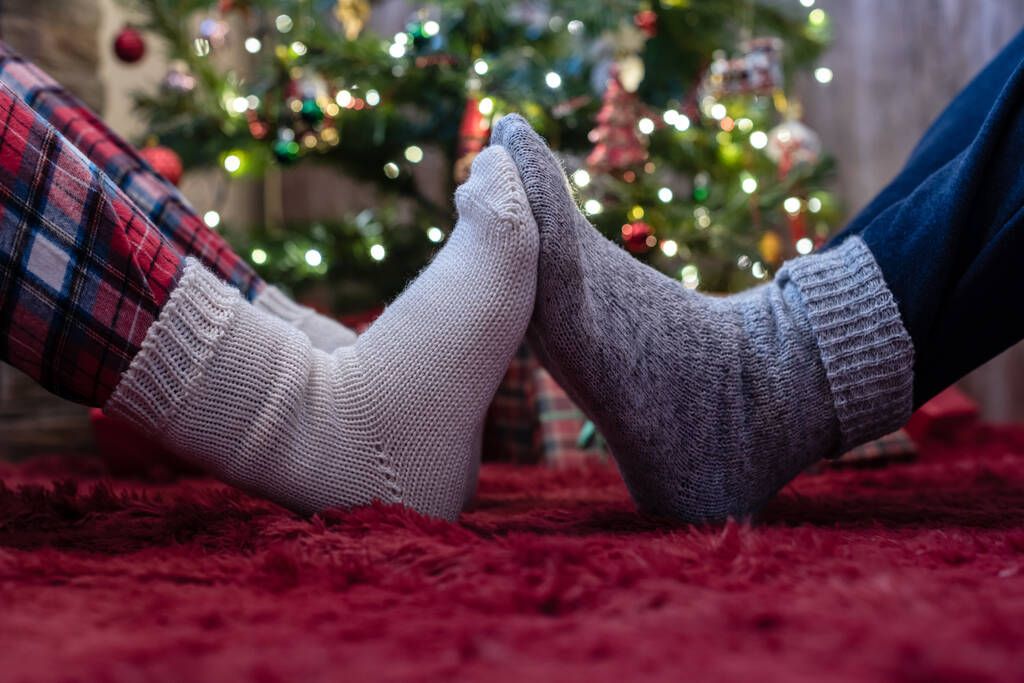 Kış çoraplarının içindeki erkek ve kadın ayakları birbirine dokunuyor. Noel ağacının yanında hediyelerle oturan adam ve kadın. Kavram - Fotoğraf, Görsel