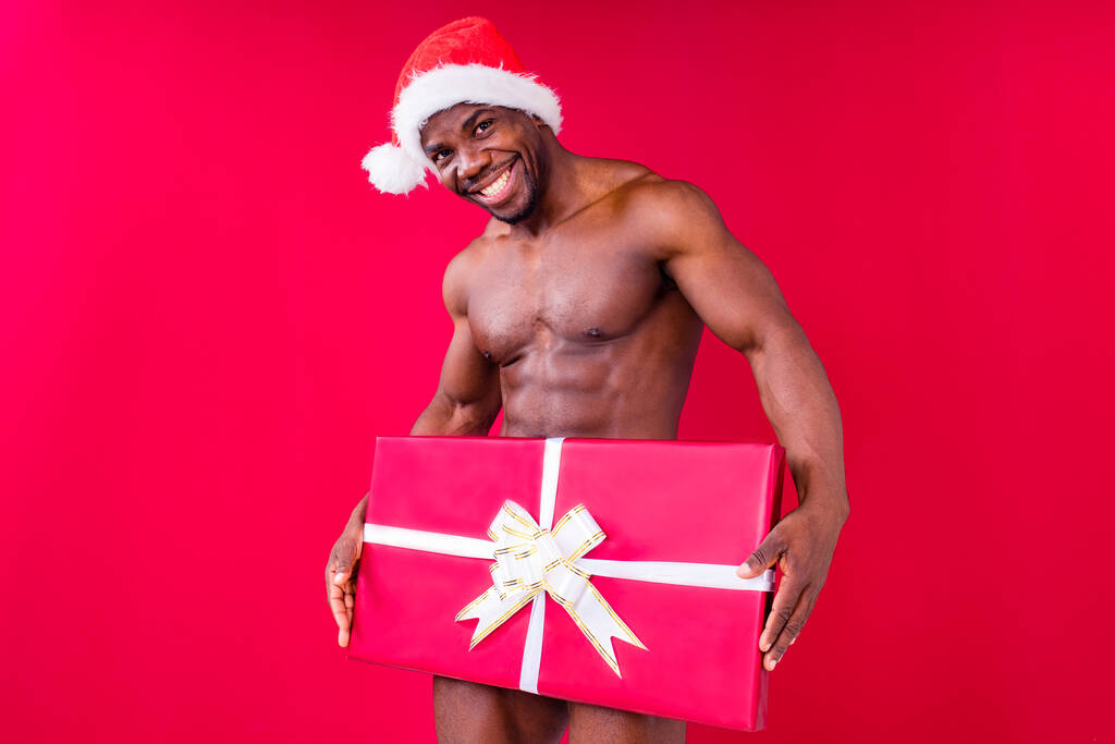 Çıplak, Afro-Amerikalı, elinde beyaz kurdeleli hediye kutusu olan bir adam kırmızı stüdyo arka planında papyon takıyor. - Fotoğraf, Görsel