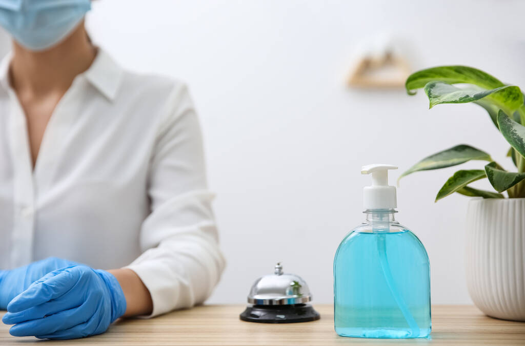 Receptionist at counter top in hotel, focus on dispenser bottle with antiseptic gel and service bell - Фото, изображение