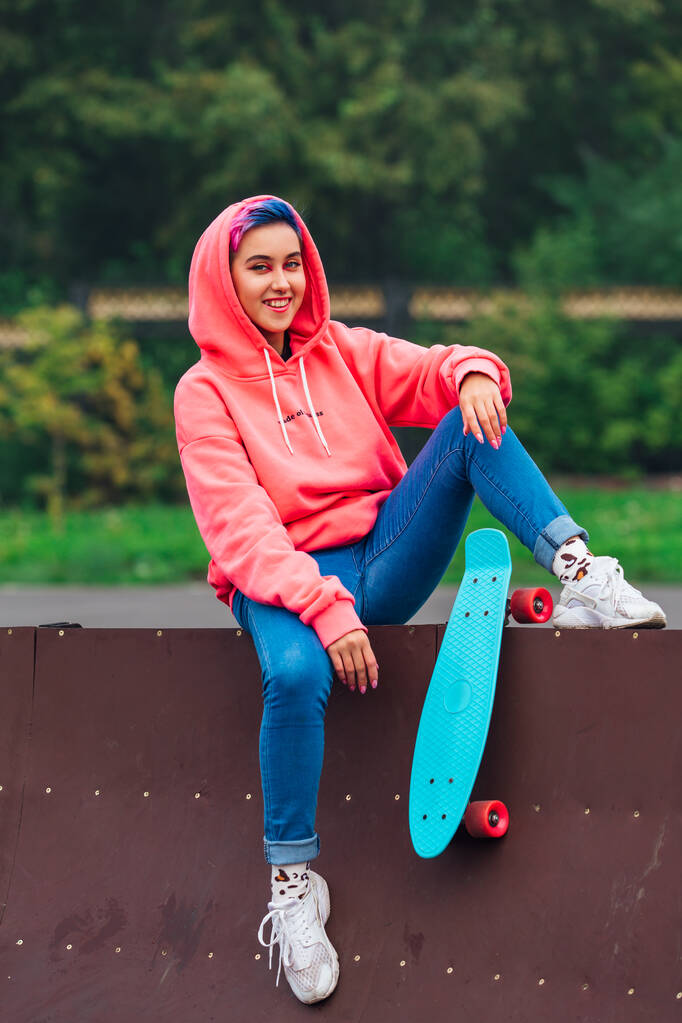 Portret van een trendy mooi jong meisje met kort gekleurd haar en neus piercing gekleed in roze hoodie en jeans zitten naast de skateboard court met haar blauwe plastic skateboard. - Foto, afbeelding