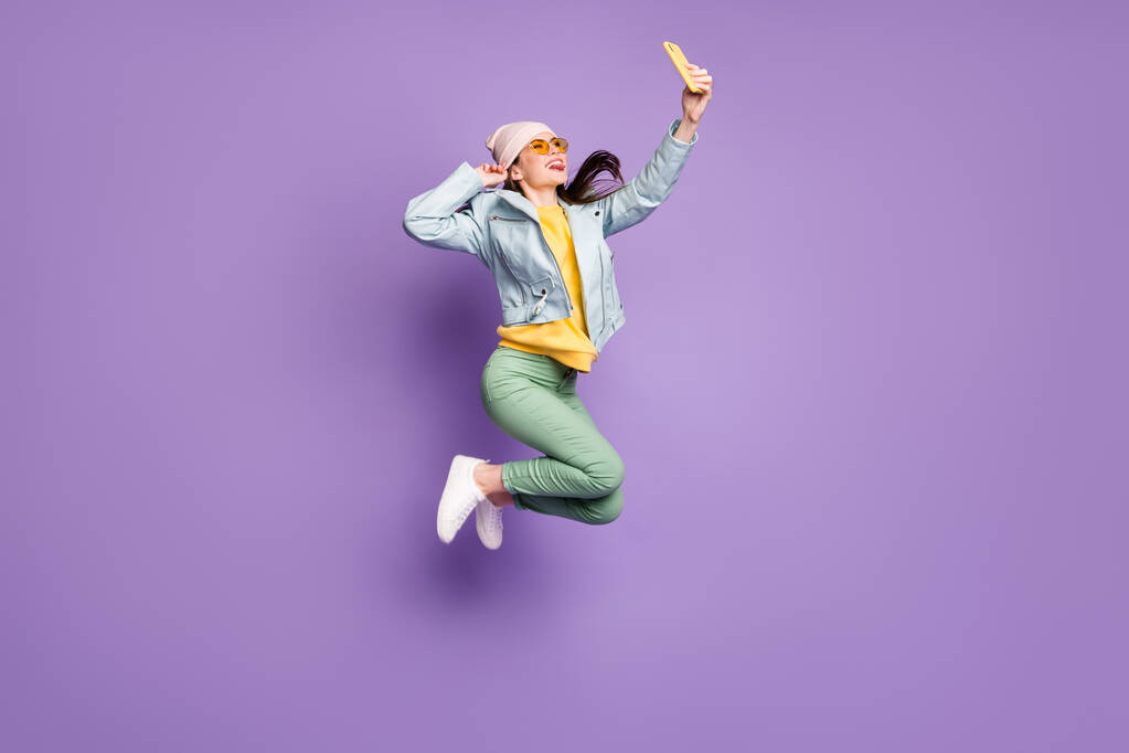 フル長さの写真面白い愚かな女の子は秋の自由時間ジャンプ作る自撮りスマートフォンショー舌アウト着用革ジャケットサングラス緑のズボン孤立紫色の背景 - 写真・画像