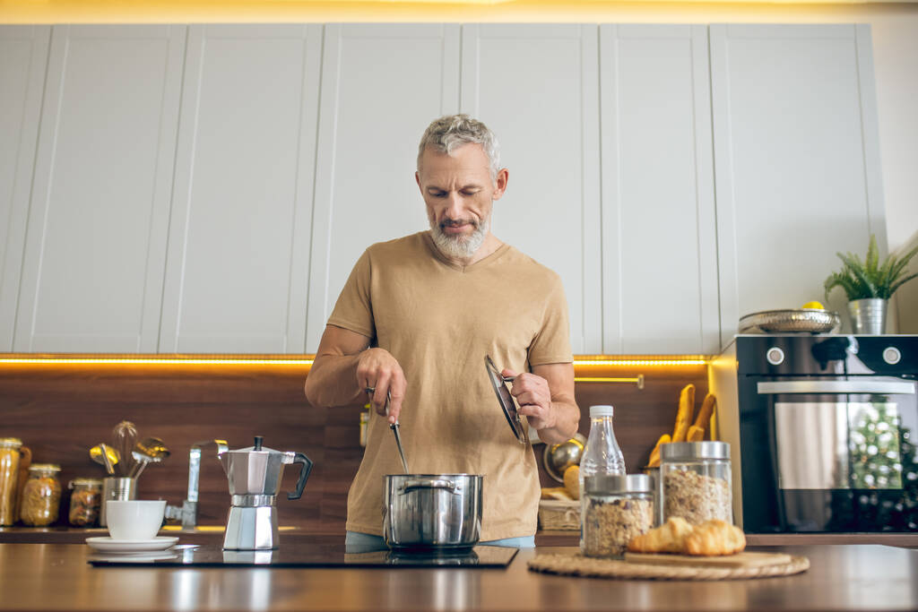 Kypsä mies beige tpaita seisoo keittiössä ja tekee aamiaista - Valokuva, kuva