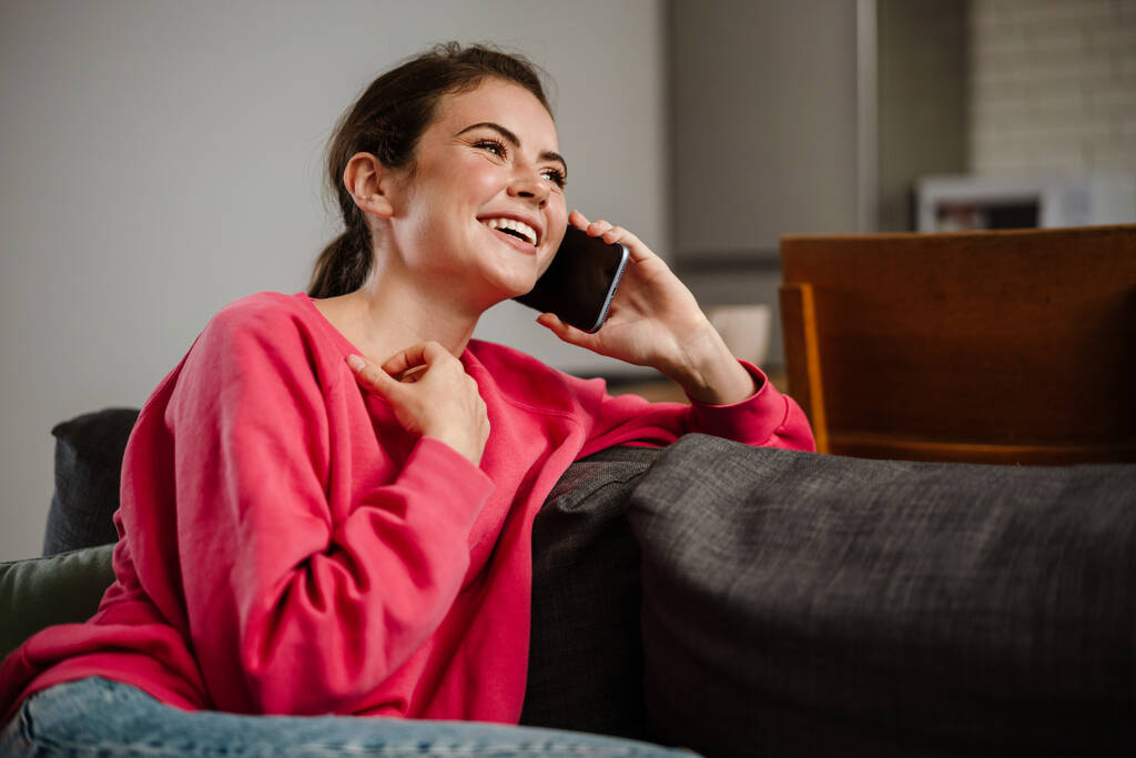 Gyönyörű örömteli lány beszél mobilon és mosolyog, miközben otthon ül a kanapén. - Fotó, kép