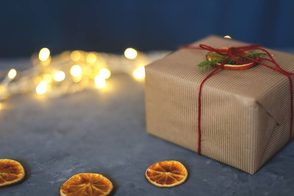 Caja de regalo hecha a mano decorada con papel artesanal con un pedazo de naranja, con luces sobre fondo gris y azul, vista superior. Feliz tarjeta de felicitación navideña. Tema de vacaciones de Navidad de invierno. Puesta plana - Foto, Imagen