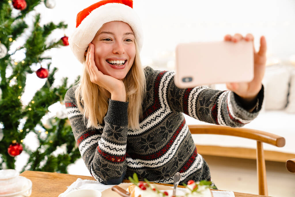 Kép egy fiatal boldog nő karácsonykor Mikulás kalap otthon csinál egy szelfi okostelefon - Fotó, kép