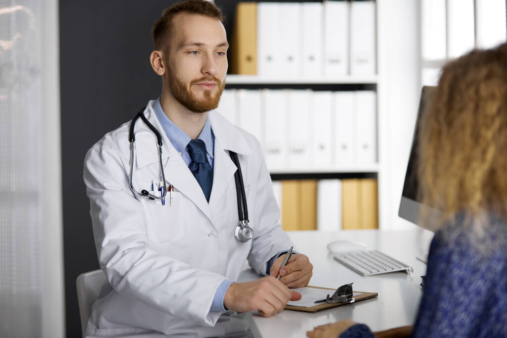 Friendly red-bearded doctor and patient woman discussing current health examination while sitting in sunny clinic. Medicine concept - Photo, Image