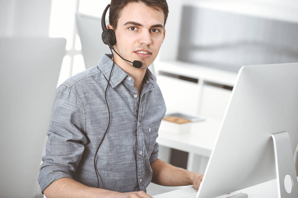 オンラインで顧客と話をしながら、ヘッドセットとコンピュータを使用してカジュアルな服装の若い男。コールセンター、ビジネスコンセプト - 写真・画像