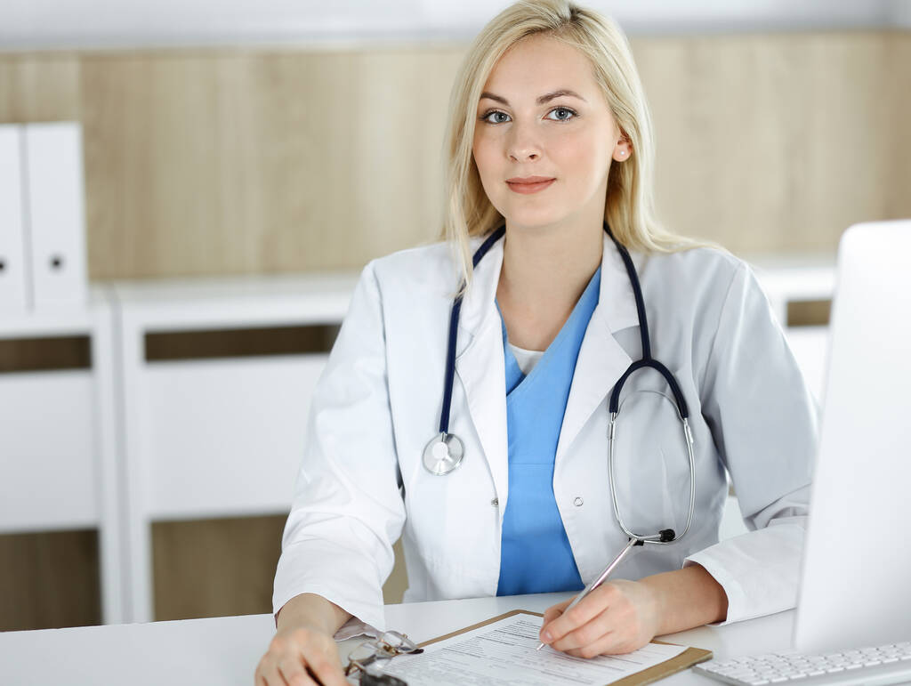 Retrato de mujer-médico en el trabajo mientras está sentado en el escritorio en la clínica. Rubia médico alegre llenando formulario médico o prescripción - Foto, Imagen