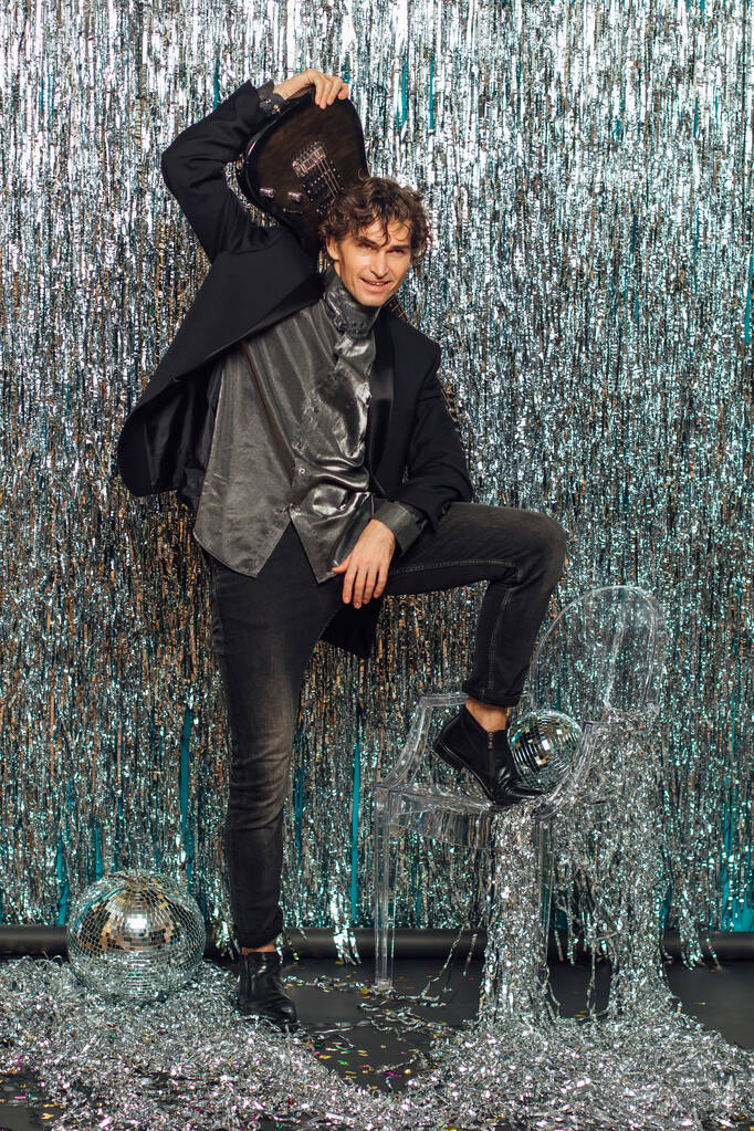 Tall attractive man holding electro guitar standing over the sparkle background with a mirror disco ball on the floor. Party concept. - Photo, Image