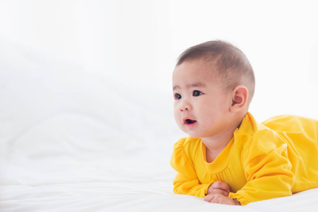 Retrato de un hermoso bebé recién nacido asiático joven propenso en la cama en casa, Feliz sonrisa del bebé lleva una camisa amarilla relajante en la habitación, Mañana familiar en casa - Foto, imagen