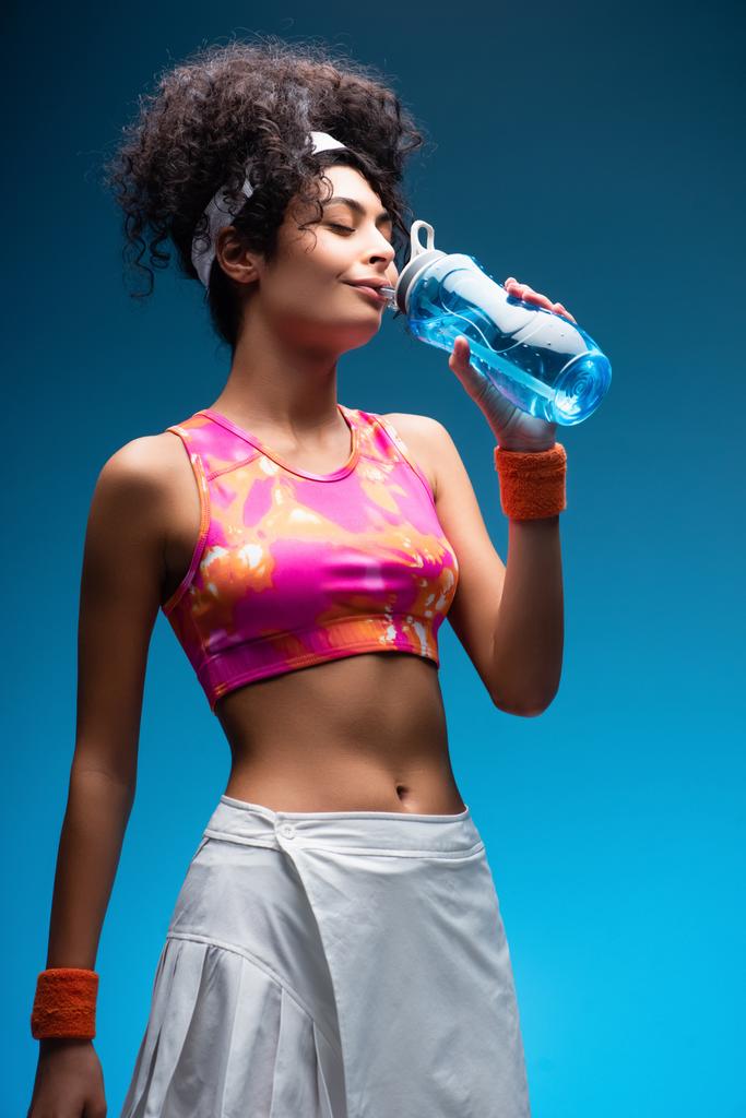 σγουρή γυναίκα σε αθλητικά ρούχα πόσιμο νερό σε μπλε - Φωτογραφία, εικόνα
