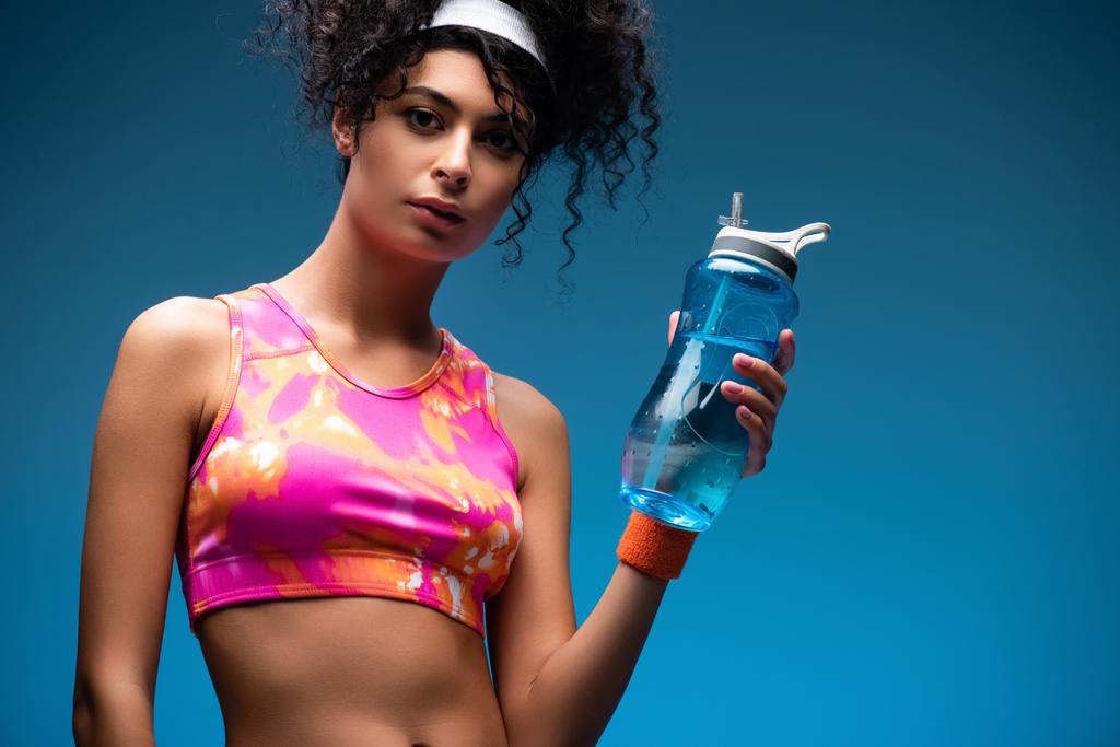 femme bouclée en vêtements de sport tenant bouteille de sport avec de l'eau sur bleu - Photo, image