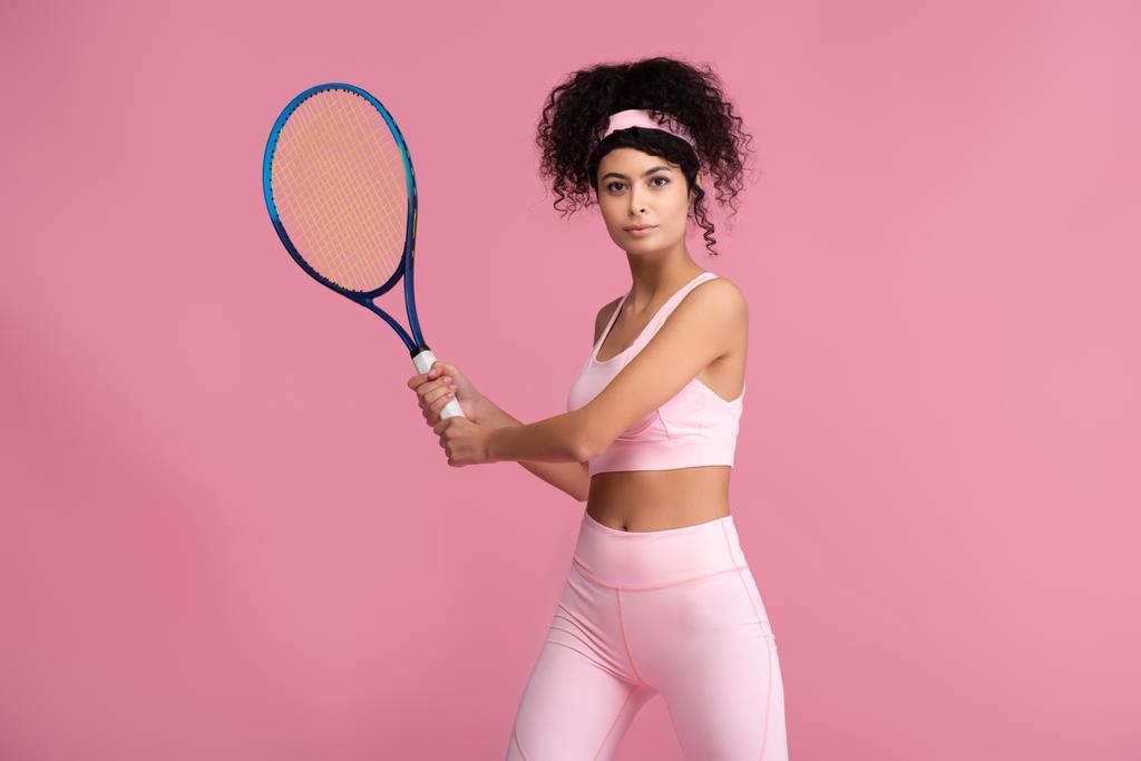 young curly sportswoman holding tennis racket while playing isolated on pink - Photo, Image