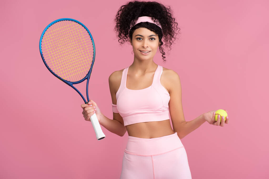 šťastná a sportovní mladá žena drží tenisovou raketu a míč izolované na růžové  - Fotografie, Obrázek